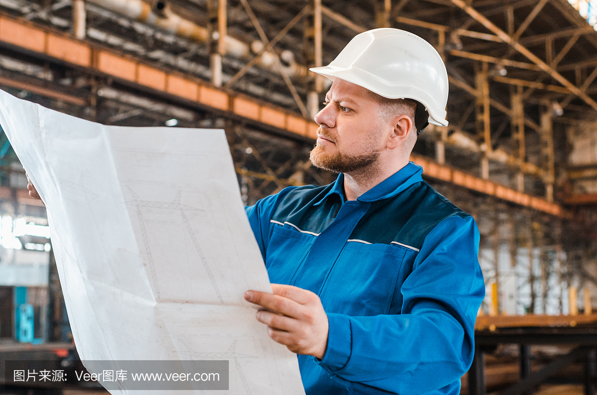 一個穿著工作服、戴著白色頭盔的工人正在檢查一個金屬工廠背景上的圖紙。建筑工人身體很好。男工程師